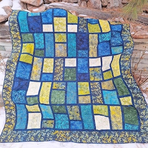 Pode incluir: Um cobertor de retalhos azul, verde e amarelo com um padrão floral. O cobertor é feito de quadrados e retângulos de diferentes tamanhos e cores. O cobertor está dobrado ao meio e está sobre uma superfície nevada.