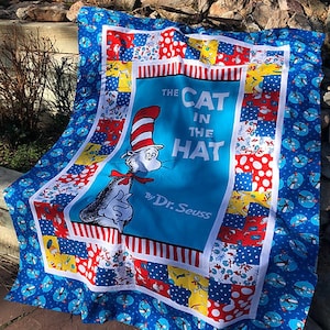 May include: A blue and white quilt with a patchwork design featuring various patterns, including polka dots, stripes, and floral prints. The center panel features a colorful illustration of the Cat in the Hat from the Dr. Seuss book.