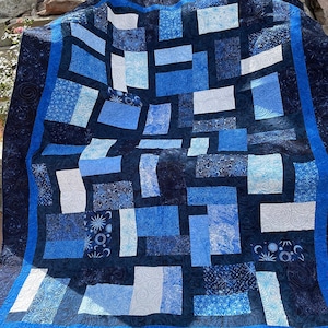 May include: A blue and white patchwork quilt with a dark blue border. The quilt is made of various shades of blue and white fabric, and the border is decorated with a swirling pattern.