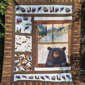 May include: A rectangular quilt featuring a brown wood-grain border and a central panel depicting a black bear looking through a window at a mountain lake scene. The quilt includes smaller square panels with bear, canoe, and camping motifs.