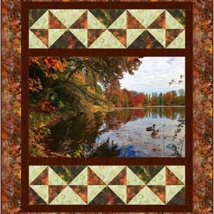 May include: A quilted wall hanging featuring a serene autumn lake scene. The central image is framed by a brown border and decorative panels with triangle patterns in shades of green, brown, and cream. The outer border has a leaf pattern.