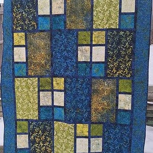 Może przedstawiać: Kołdra w kolorze niebieskim, zielonym i żółtym z geometrycznym wzorem. Kołdra składa się z kwadratów i prostokątów, a kolory są ułożone w powtarzający się wzór.