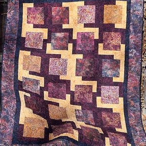 May include: A handmade quilt with a geometric pattern. The quilt features squares in shades of purple, gold, and patterned fabrics. The border is a dark blue with a swirling design. The quilt is displayed outdoors.