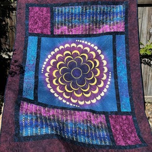 May include: A quilt with a dark blue background and a large, circular design of gold crescent moons. The design is surrounded by a border of purple and black fabric. The quilt is made up of several different pieces of fabric, each with a unique pattern.