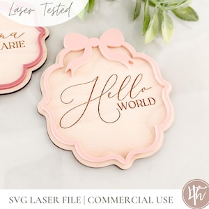 Puede incluir: Un letrero de madera con un lazo rosa y el texto "Hello World" grabado en &eacute;l. El letrero est&aacute; cortado con l&aacute;ser y tiene un borde festoneado.