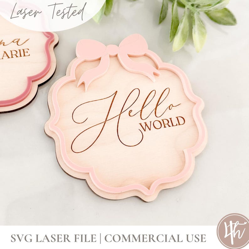 Puede incluir: Un letrero de madera con un lazo rosa y el texto "Hello World" grabado en &eacute;l. El letrero est&aacute; cortado con l&aacute;ser y tiene un borde festoneado.