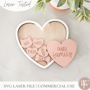 May include: A white heart-shaped frame with a wooden background holds smaller pink hearts with names like "Leo", "Miles", "Tyler", "Laura", and "Lucy". A larger pink heart with the text "our family" is partially visible outside the frame.
