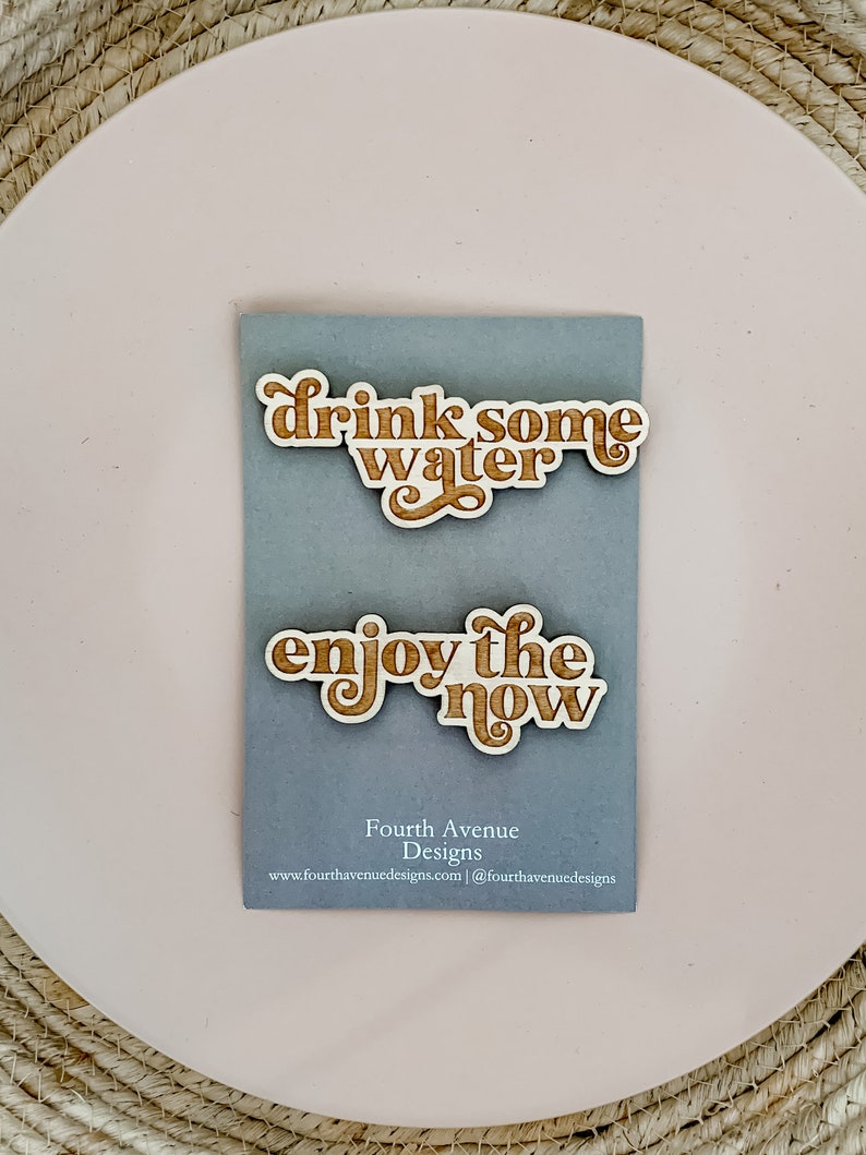 May include: Two wooden signs with a light brown wood grain and white text. The top sign says "drink some water" and the bottom sign says "enjoy the now". The signs are on a gray background with the text "Fourth Avenue Designs" and a website address.