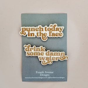 May include: Two wooden signs with motivational phrases in a retro font. The top sign reads "punch today in the face" and the bottom sign reads "drink some damn water".