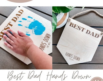 Może przedstawiać: Drewniany znak z napisem "Best Dad Hands Down" w brązowych literach. Znak jest ozdobiony niebieskim odciskiem dłoni i jest gotowy do spersonalizowania.