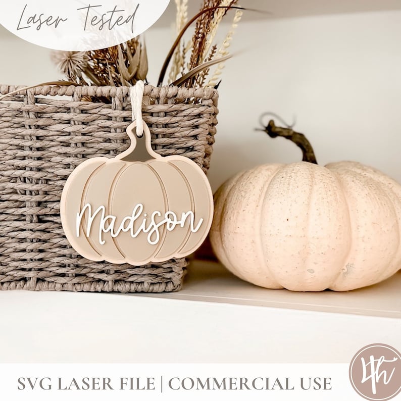 May include: A light brown pumpkin-shaped wooden ornament with the name "Madison" written in white. The ornament is hanging from a string and is sitting on a shelf next to a wicker basket and a white pumpkin.