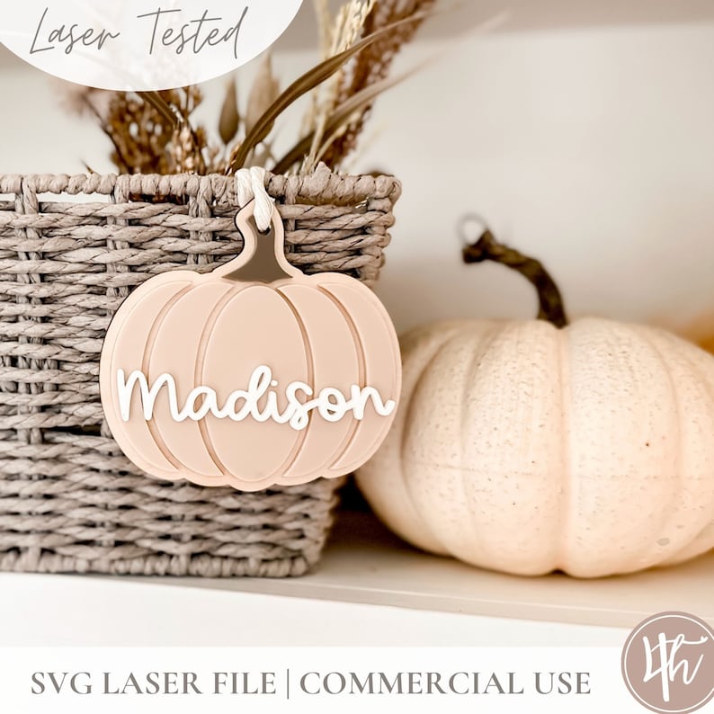 May include: A light pink pumpkin-shaped ornament with the name "Madison" written in white. The ornament is hanging from a wicker basket.