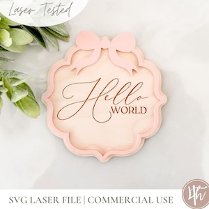 Puede incluir: Un letrero de madera rosa claro con un lazo decorativo en la parte superior y las palabras "Hello World" grabadas en el centro.