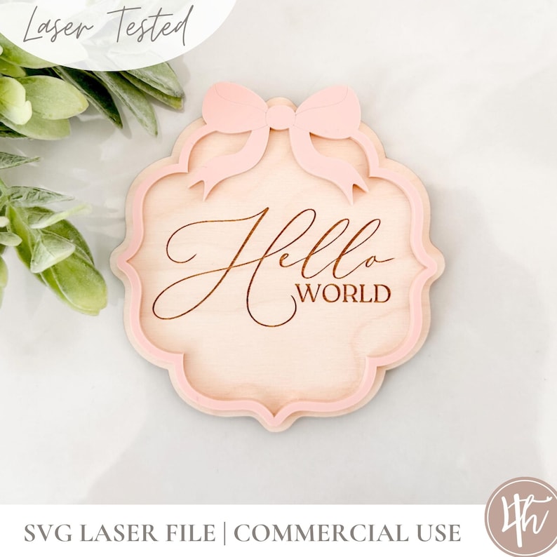 Puede incluir: Un letrero de madera rosa claro con un lazo decorativo en la parte superior y las palabras "Hello World" grabadas en el centro.
