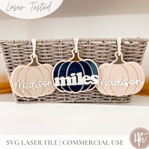 May include: Three pumpkin-shaped ornaments with the names "Madison", "miles", and "Madison" written on them. The pumpkins are in a wicker basket. The pumpkins are pink and blue.