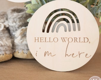 May include: A wooden circle with a rainbow cutout and the text "HELLO WORLD, i'm here" in a handwritten font. The circle is sitting on a brown surface with a pair of knitted baby booties.