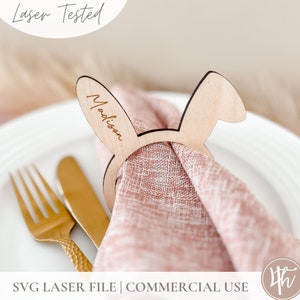 Può includere: Un anello porta tovagliolo di legno a forma di coniglio con il nome "Madison" inciso. L'anello tiene un tovagliolo rosa e si trova su un piatto bianco.