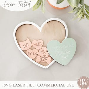 May include: A white heart-shaped frame with a wooden interior holds a collection of smaller pink hearts with names inscribed on them, including "Leo", "Miles", "Tyler", "Laura", and "Lucy". A green heart with the text "Loved with all Nana's heart" is placed on the right side of the frame.