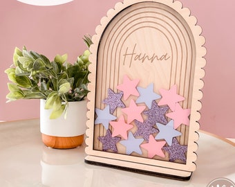 Pode incluir: Um letreiro de madeira com forma de arco festoneado, apresentando um design de arco-íris e o nome "Hanna" inscrito. O letreiro tem uma ranura para inserir pequenas decorações em forma de estrela, com estrelas rosa e roxas visíveis.