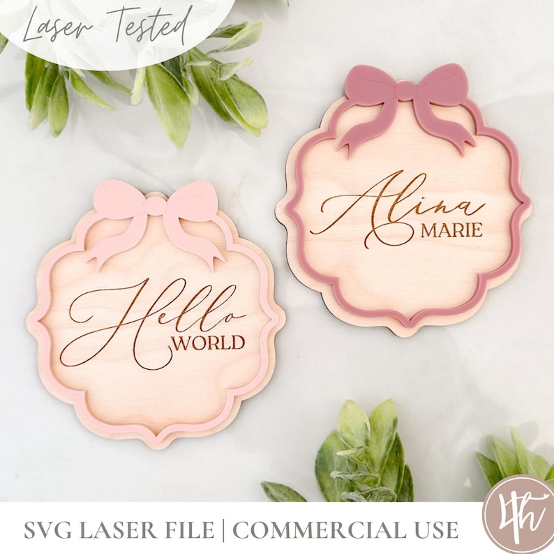 Puede incluir: Dos letreros de madera con lazos rosas y texto grabado. El primer letrero dice "Hello World" y el segundo letrero dice "Alina Marie".