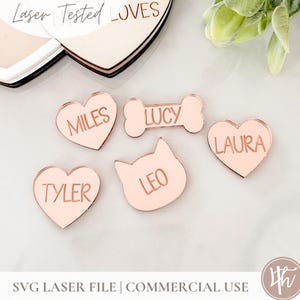 May include: Five pink acrylic pet name tags with rose gold lettering. The tags are shaped like hearts, a bone, and a cat head. The names on the tags are Miles, Lucy, Laura, Tyler, and Leo.