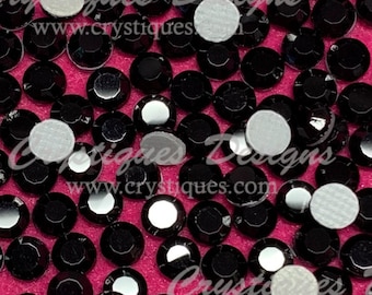 May include: A close-up image of black rhinestones with adhesive backing. The rhinestones are scattered on a pink surface.