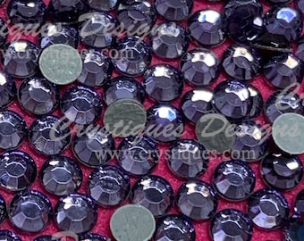 May include: A close-up image of a large quantity of small, round, iridescent purple rhinestones. The rhinestones are scattered on a pink surface.