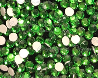 May include: A close-up image of a large quantity of small, round, green rhinestones. The rhinestones have a shiny, faceted surface and are scattered on a black background.