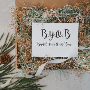 May include: A white card with the text "B.y.o.B. Build Your Own Box" inside a cardboard box filled with brown and blue shredded paper.