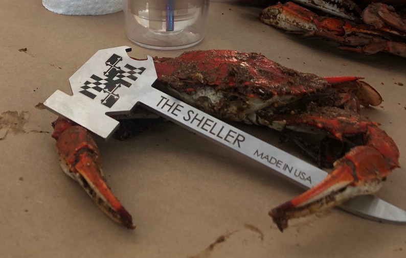 Może przedstawiać: Srebrny rozdrabniacz do krab&oacute;w z czarno-białym wzorem w kratkę i tekstem "THE SHELLER MADE IN USA". Narzędzie leży na brązowej powierzchni obok gotowanego kraba.
