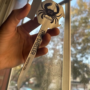Może przedstawiać: Srebrny otwieracz do butelek w kształcie kraba z napisem "The Sheller" i "Made in USA". Krab ma napis "Shore" i "Maryland".