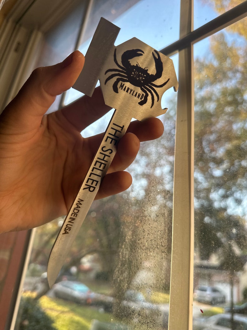 Może przedstawiać: Srebrny otwieracz do butelek w kształcie kraba z napisem "The Sheller" i "Made in USA". Krab ma napis "Shore" i "Maryland".