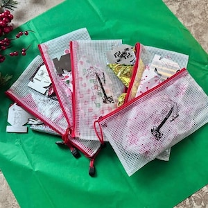 May include: Several white mesh zipper pouches with red trim, containing various items, are arranged on a green surface. One pouch features the text "The Shelter" and a graphic. Some pouches have red and white patterns.
