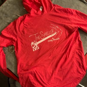 May include: A red hooded sweatshirt with long sleeves. The white text on the front reads "The Sheller" with a graphic of a crab claw and the text "by Shelby Blondell". The sweatshirt is made of a soft material.