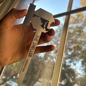 Może przedstawiać: Srebrny otwieracz do butelek w kształcie stanu Maryland z napisem "The Sheller Made in USA" na uchwycie.