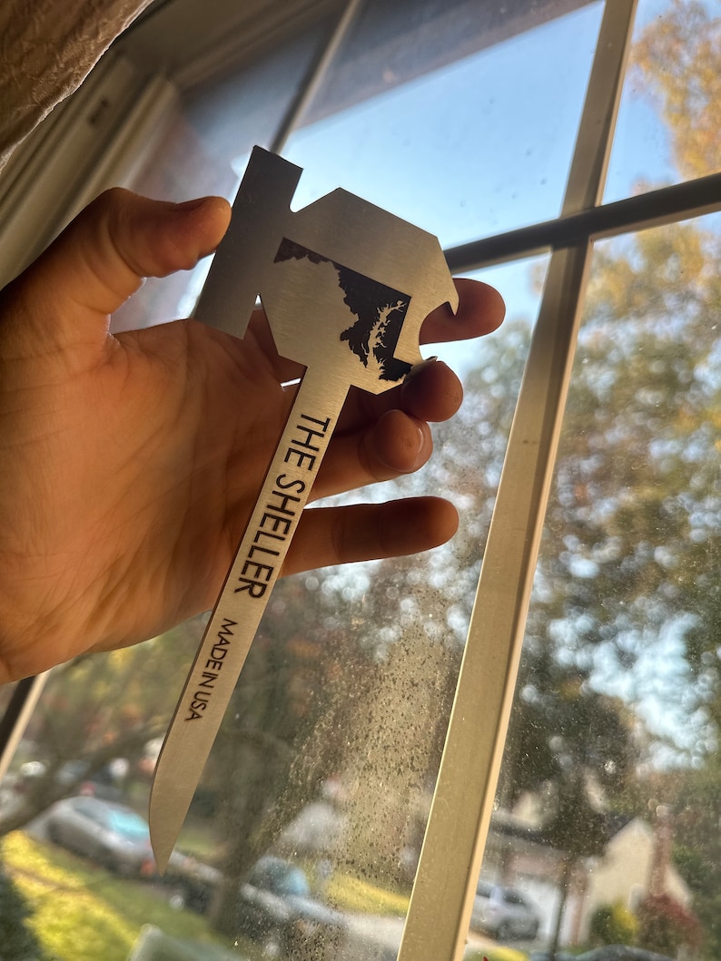 Może przedstawiać: Srebrny otwieracz do butelek w kształcie stanu Maryland z napisem "The Sheller Made in USA" na uchwycie.