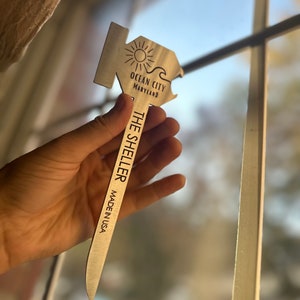 Może przedstawiać: Srebrny otwieracz do butelek w kształcie domu z motywem słońca i fali. Na otwieraczu wygrawerowano tekst "Ocean City Maryland" i "The Sheller Made in USA".