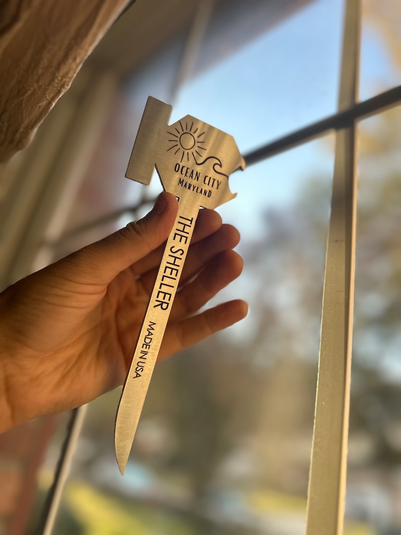 Może przedstawiać: Srebrny otwieracz do butelek w kształcie domu z motywem słońca i fali. Na otwieraczu wygrawerowano tekst "Ocean City Maryland" i "The Sheller Made in USA".