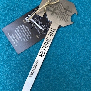 May include: A silver metal bottle opener with a pointed end and a bottle opener on the top. The bottle opener has the text "The Sheller Made in USA" printed on it.
