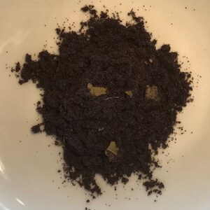 Pode incluir: Close-up de um monte de pó marrom escuro com pequenos pedaços verdes misturados. O pó está sobre uma superfície branca.