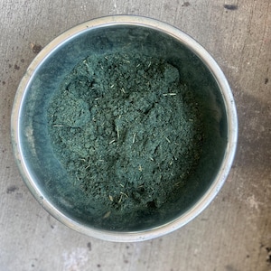 May include: A metal bowl filled with a dark green powder. The powder is finely ground and appears to be a type of herb or spice.