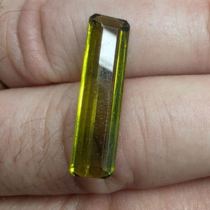 May include: A rectangular, faceted gemstone with a vibrant olive-green hue and a translucent, glassy appearance. The stone features a long, slender shape with sharp, clean edges and a polished surface that reflects light across its elongated facets.