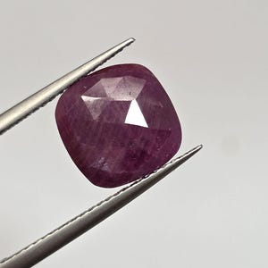 May include: A faceted, cushion-cut ruby gemstone held by silver tweezers. The ruby is a deep, saturated red-purple color, with multiple facets catching the light. The gemstone is a square shape with rounded corners.