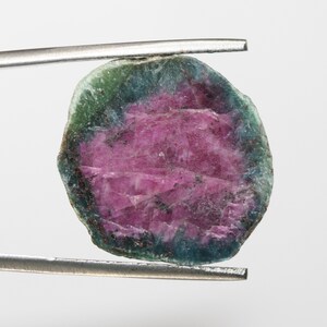 May include: A round, green and pink gemstone with a rough, natural finish. The gemstone is held in place by a pair of tweezers.