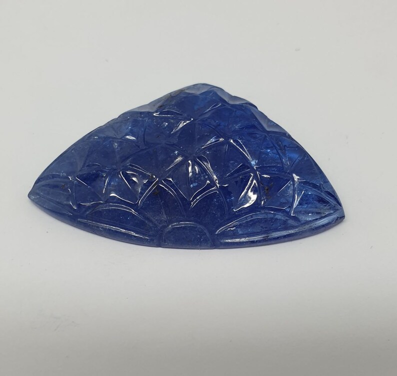 May include: A carved blue gemstone with a fan-shaped design. The gemstone is a triangular shape with a smooth, polished surface.