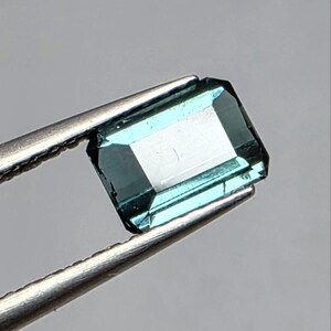 Pode incluir: Uma pedra preciosa retangular com corte esmeralda e um tom azul-esverdeado profundo. A pedra apresenta superfícies facetadas que refletem a luz, criando uma aparência clara e transparente. É segurada por uma pinça de metal prateado.