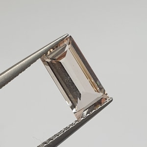 May include: A light brown rectangular gemstone with a faceted cut, held in tweezers.