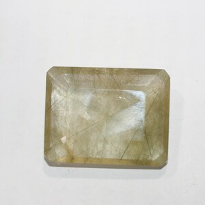 Puede incluir: Una gema rectangular de color marrón claro con una red de inclusiones oscuras en forma de cabello. La gema tiene un corte facetado con una superficie pulida, que refleja la luz. La forma general es un rectángulo con esquinas cortadas.