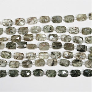 Pode incluir: Um fio de contas retangulares facetadas verdes e transparentes com um brilho prateado. As contas são enfiadas em um fio branco.