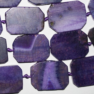 Peut inclure: Un collier de perles d'agate violette avec une finition polie et facetée. Les perles sont de forme rectangulaire et présentent un motif naturel et organique.